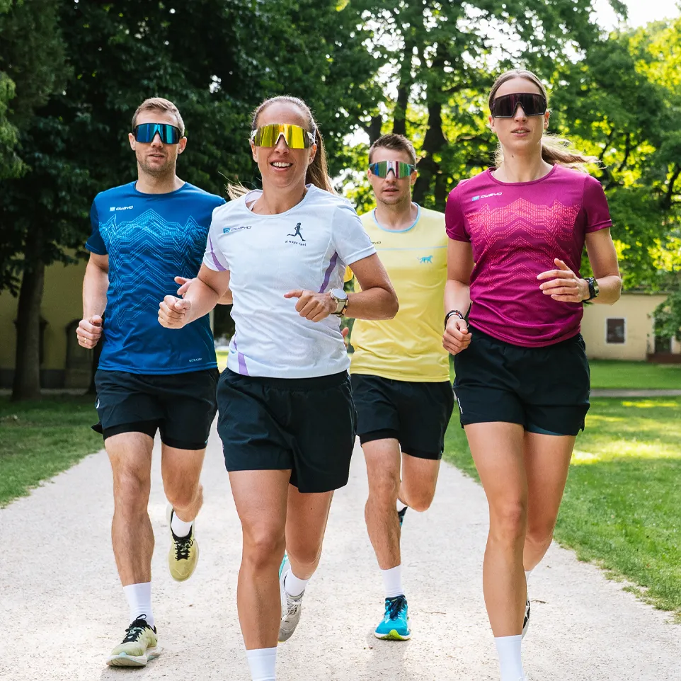 Four runners in colorful sports shirts and shorts jog on a path in a park.